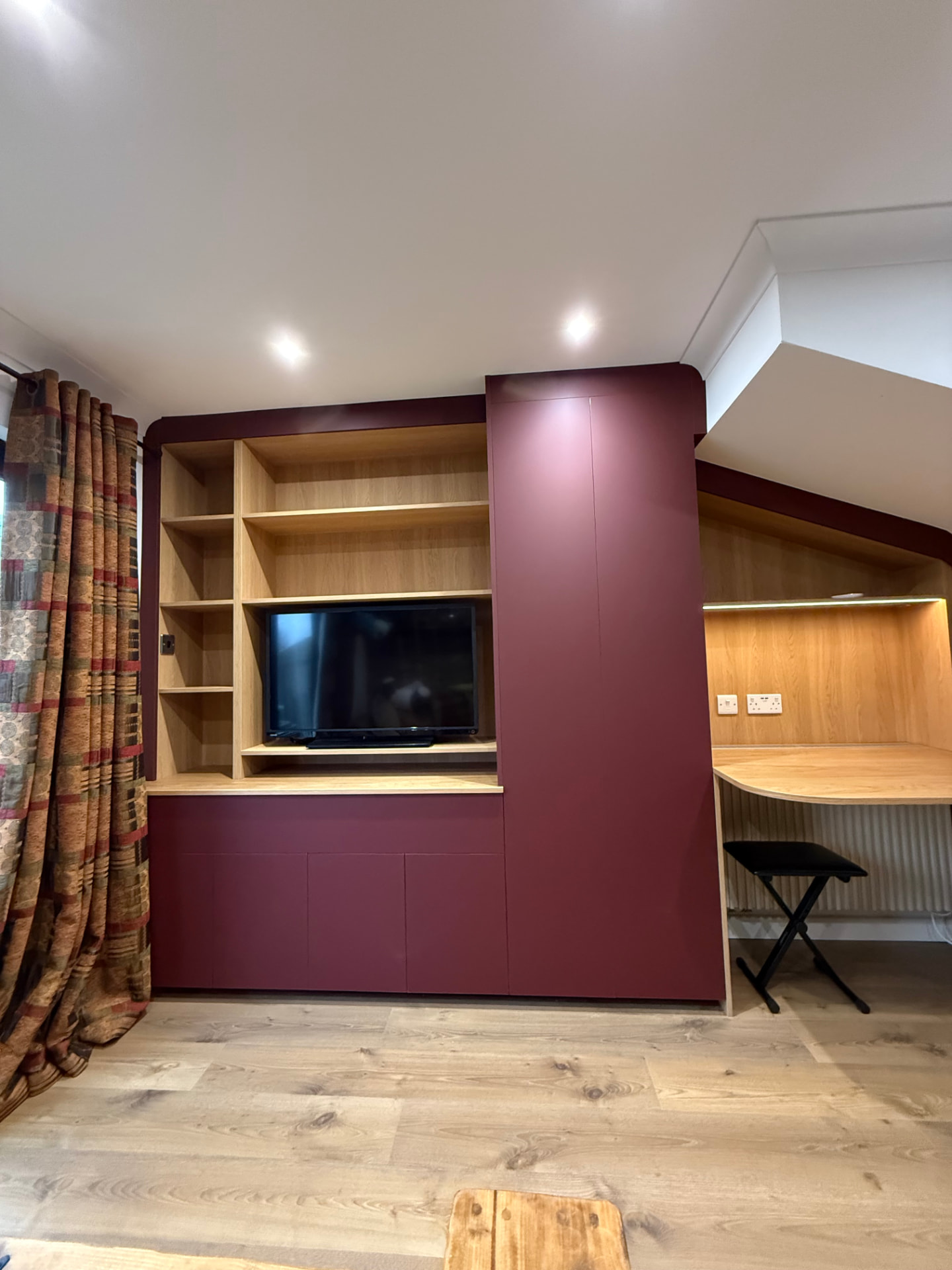 Bespoke burgundy media wall with oak shelving and integrated home office desk under a sloped ceiling