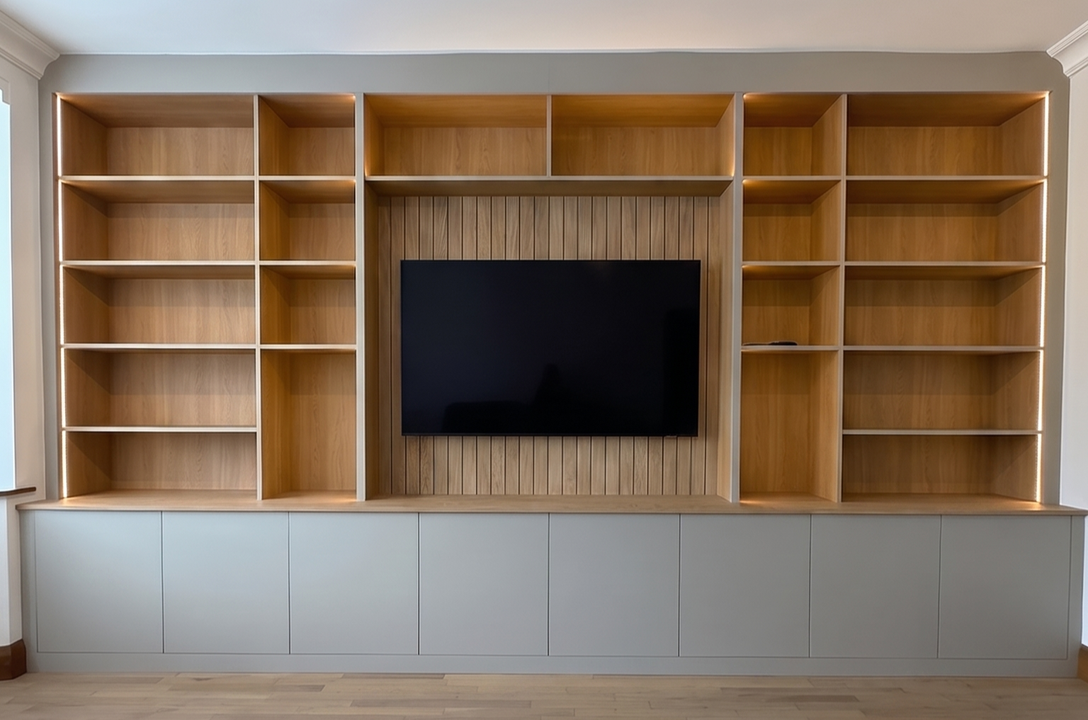 Light grey bespoke media wall with oak-slat TV alcove and illuminated shelving